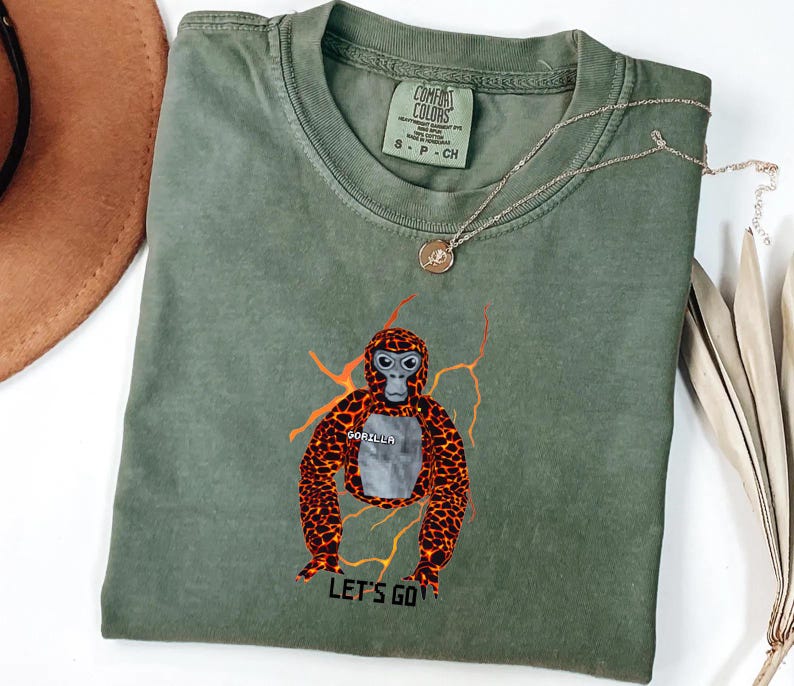 May include: An olive green t-shirt featuring a graphic of a gorilla with orange and brown fur and the text "LET'S GO". The shirt also has the text "GORILLA" on the chest. A gold necklace with a pendant is draped across the shirt.