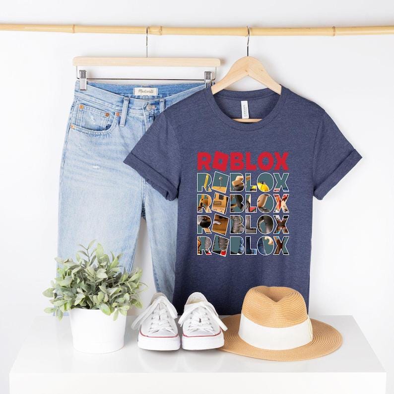 May include: Navy blue t-shirt with the word "ROBLOX" in red block letters. The word "ROBLOX" is repeated multiple times in a stacked design. Light blue denim jeans, white sneakers, and a straw hat are also in the image.