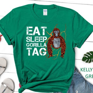 May include: Kelly green t-shirt with the text "EAT SLEEP GORILLA TAG" in white block letters. A cartoon gorilla with orange and brown fur is featured on the right side. The shirt is a Gildan brand.