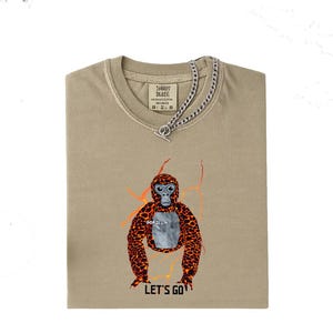 May include: Tan t-shirt with a graphic of an orangutan with orange and brown spots and the text "LET'S GO". The shirt has a silver chain necklace. The shirt is folded.