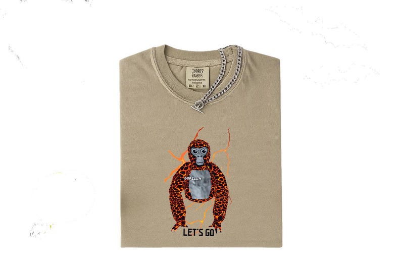 May include: Tan t-shirt with a graphic of an orangutan with orange and brown spots and the text "LET'S GO". The shirt has a silver chain necklace. The shirt is folded.