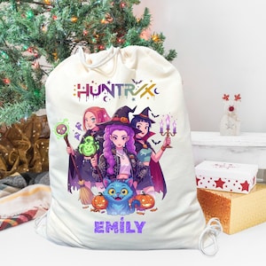 May include: A large, off-white drawstring bag featuring a colorful illustration of three witches, a cat, and pumpkins. The bag has the text "HUNTRIX" and "EMILY" printed on it. A Christmas tree is in the background.