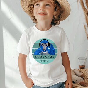 May include: White t-shirt featuring a blue monkey wearing headphones, set within a teal and white circular design. The text "COME AT ME BRUH" is below the monkey. The shirt is worn by a child.