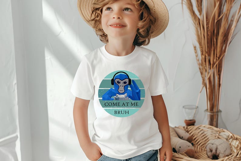 May include: White t-shirt featuring a blue monkey wearing headphones, set within a teal and white circular design. The text "COME AT ME BRUH" is below the monkey. The shirt is worn by a child.