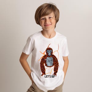 May include: White t-shirt featuring a cartoon gorilla with orange and brown fur, blue eyes, and the word "GORILLA" on its chest. The shirt also has the text "LET'S GO" at the bottom. The gorilla design is surrounded by orange lightning bolt graphics.