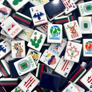 K&ouml;nnte beinhalten: Eine Sammlung wei&szlig;er Mahjong-Spielsteine mit verschiedenen bunten Illustrationen, darunter ein Gepard, ein Hirsch, ein Kaktus, ein Adler, ein Ahornblatt und Stadiongrafiken. Einige Steine zeigen den Text JOKER, SOUTH, NORTH oder CRAK in Rot oder Blau.