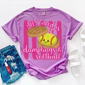 May include: A lilac-colored t-shirt with the text "All I need is dumplings & softball." The graphic features a cartoon softball in a bamboo steamer basket. The shirt is paired with denim shorts and white sandals.