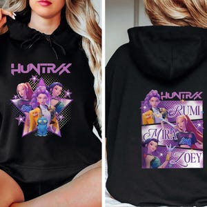 May include: Black hoodie featuring a graphic of the word "HUNTRAX" above a star-shaped design with three animated characters. The back of the hoodie displays the word "HUNTRAX" and the names "RUMI", "MIRA", and "ZOEY".