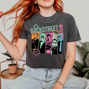 May include: A charcoal gray t-shirt featuring the text BACKSTREET BOYS in a neon font above four rectangular panels. Each panel contains a portrait of a band member in shades of yellow, green, pink, and blue, with geometric shapes surrounding them.