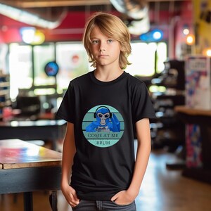 May include: Black t-shirt featuring a blue cartoon monkey wearing headphones and holding a game controller. The design is set within a circle with blue horizontal stripes and the text "COME AT ME BRUH".