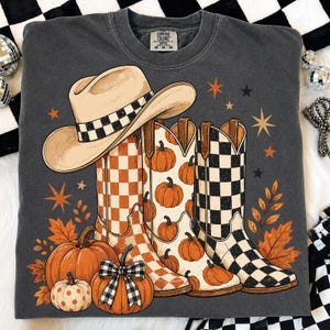 May include: A charcoal gray crewneck sweatshirt featuring a graphic print of a tan cowboy hat, three cowboy boots with orange checkered, pumpkin, and black checkered patterns, orange pumpkins, and orange stars on a dark gray fabric background.