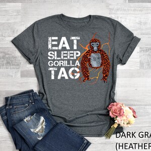 May include: Dark gray heather t-shirt with the words "EAT SLEEP GORILLA TAG" in white block letters. A cartoon orangutan with orange and brown fur is in the center. The shirt is paired with blue jeans.