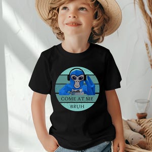 May include: Black t-shirt featuring a blue cartoon monkey wearing headphones and holding a game controller. The text "COME AT ME BRUH" is printed below the monkey in white and blue. The shirt is worn by a child.