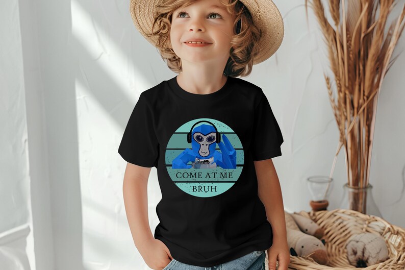 May include: Black t-shirt featuring a blue cartoon monkey wearing headphones and holding a game controller. The text "COME AT ME BRUH" is printed below the monkey in white and blue. The shirt is worn by a child.