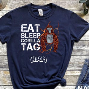 May include: Navy blue t-shirt with the text "EAT SLEEP GORILLA TAG LIAM" in white. A cartoon gorilla with orange and brown fur is featured on the shirt. The gorilla has a gray face and is surrounded by yellow lightning bolts.