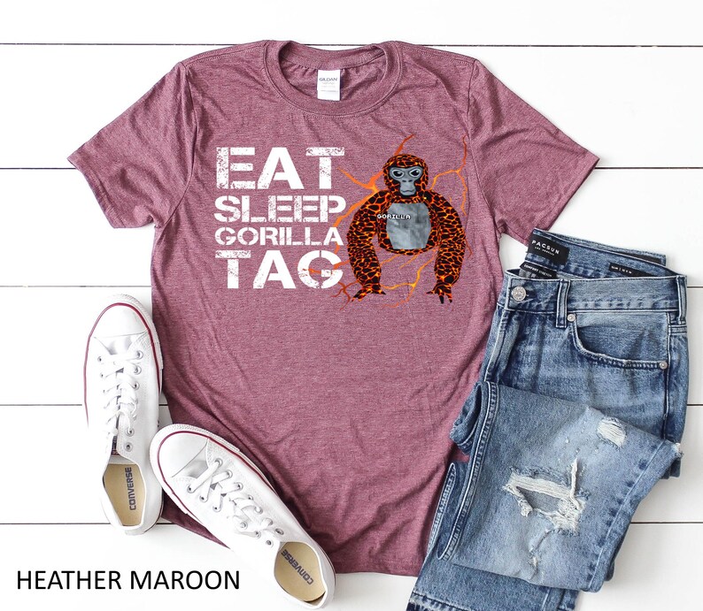 May include: Heather maroon t-shirt with the text "EAT SLEEP GORILLA TAG" in white. A cartoon gorilla with orange and brown fur is featured on the right side. The shirt is paired with blue jeans and white sneakers.