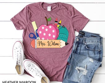 May include: Heather maroon t-shirt with a school-themed graphic. The graphic includes a pink apple with white flowers, a green backpack, crayons, scissors, and a pencil with the text "Mrs. Wilson". White sneakers and blue jeans are also in the image.