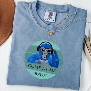 May include: A light blue t-shirt with a graphic of a blue monkey wearing headphones and the text "COME AT ME BRUH". The shirt has a crew neck and short sleeves. A gold necklace is draped across the shirt.