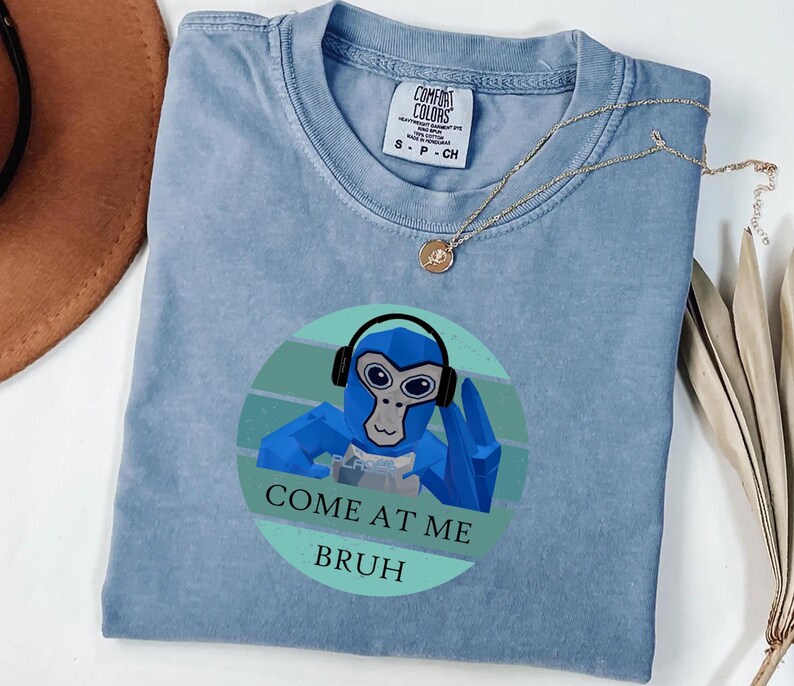 May include: A light blue t-shirt with a graphic of a blue monkey wearing headphones and the text "COME AT ME BRUH". The shirt has a crew neck and short sleeves. A gold necklace is draped across the shirt.