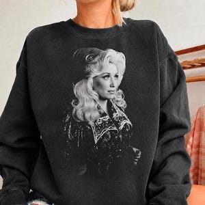 May include: A charcoal black crewneck sweatshirt featuring a large, centered black and white graphic print of a blonde woman with voluminous hair wearing a decorative jacket with intricate detailing and a dark hat.
