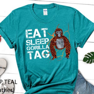 May include: Deep teal heather t-shirt with the text "EAT SLEEP GORILLA TAG" in white. A cartoon gorilla with orange and brown fur is featured on the front. Red sunglasses, silver sandals, and a green leaf keychain are also visible.