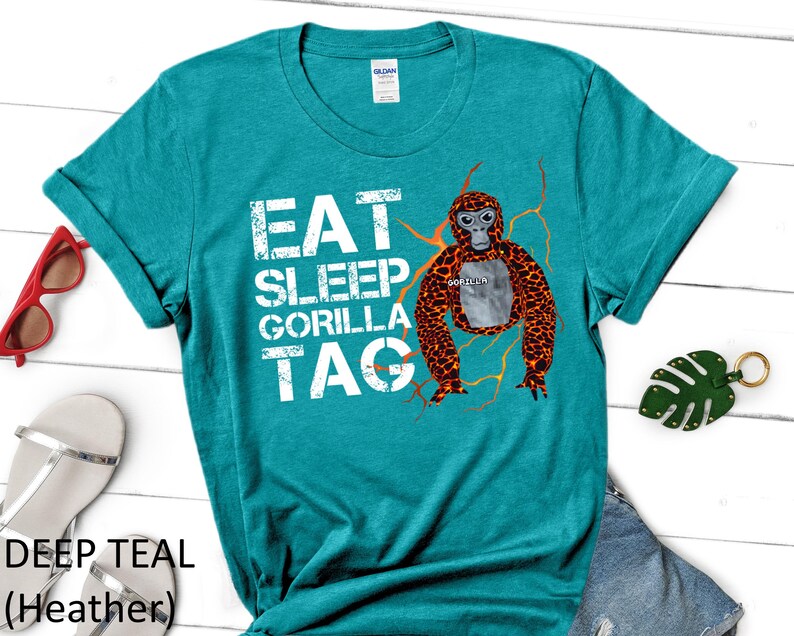May include: Deep teal heather t-shirt with the text "EAT SLEEP GORILLA TAG" in white. A cartoon gorilla with orange and brown fur is featured on the front. Red sunglasses, silver sandals, and a green leaf keychain are also visible.