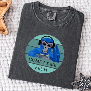 May include: Dark gray Comfort Colors t-shirt featuring a blue cartoon monkey wearing headphones. The monkey is inside a teal circle with the text "COME AT ME BRUH". The shirt is laid flat on a white surface.