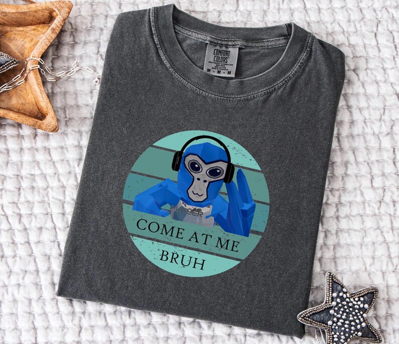 May include: Dark gray Comfort Colors t-shirt featuring a blue cartoon monkey wearing headphones. The monkey is inside a teal circle with the text "COME AT ME BRUH". The shirt is laid flat on a white surface.