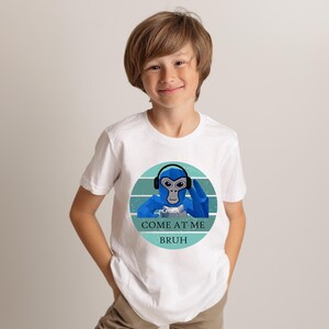 May include: White t-shirt featuring a blue cartoon monkey wearing headphones and a blue hand gesture. The text "COME AT ME BRUH" is below the monkey graphic. The shirt is being worn by a young person.