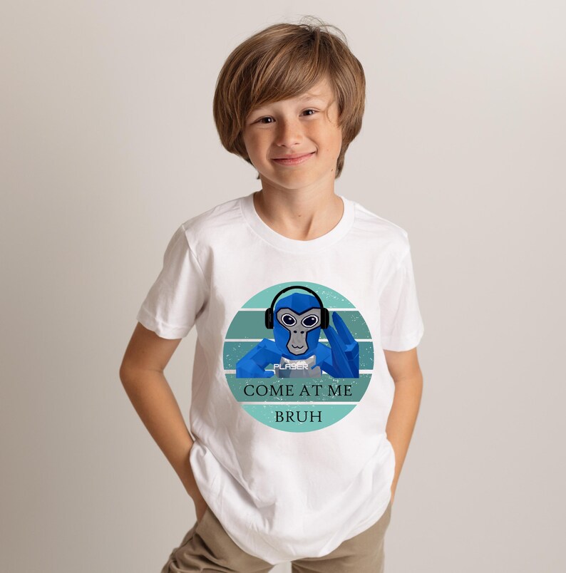 May include: White t-shirt featuring a blue cartoon monkey wearing headphones and a blue hand gesture. The text "COME AT ME BRUH" is below the monkey graphic. The shirt is being worn by a young person.