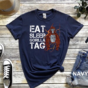 May include: Navy blue t-shirt with the text "EAT SLEEP GORILLA TAG" in white block letters. A cartoon gorilla with orange and brown fur is on the right side of the shirt. The word "GORILLA" is written on the gorilla's chest.