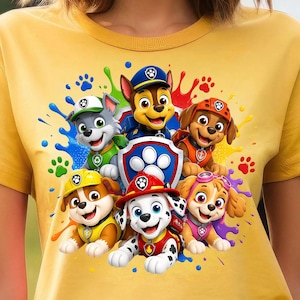 May include: A mustard yellow t-shirt featuring a colorful graphic print of six Paw Patrol characters, including Chase, Marshall, Rubble, Skye, Rocky, and Zuma, arranged around a central blue shield logo with a white paw print and paint splatters.