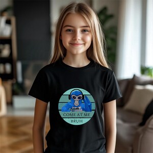 May include: Black t-shirt featuring a blue cartoon monkey wearing headphones and holding a game controller. The design is set within a teal circle with the text "COME AT ME BRUH". The shirt is worn by a person.