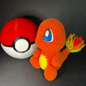 May include: An orange crochet Charmander plush toy with blue eyes and a yellow belly, next to a red and white Poké Ball. The Charmander has a colorful tail and is set against a black background.