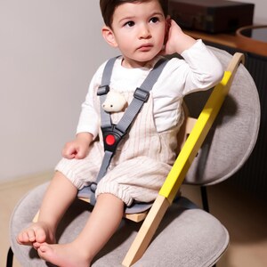 May include: A child is seated in a gray chair with a wooden high chair attachment. The high chair has a yellow accent and a gray safety harness with a red buckle. The child is wearing a white long-sleeve shirt and striped overalls.