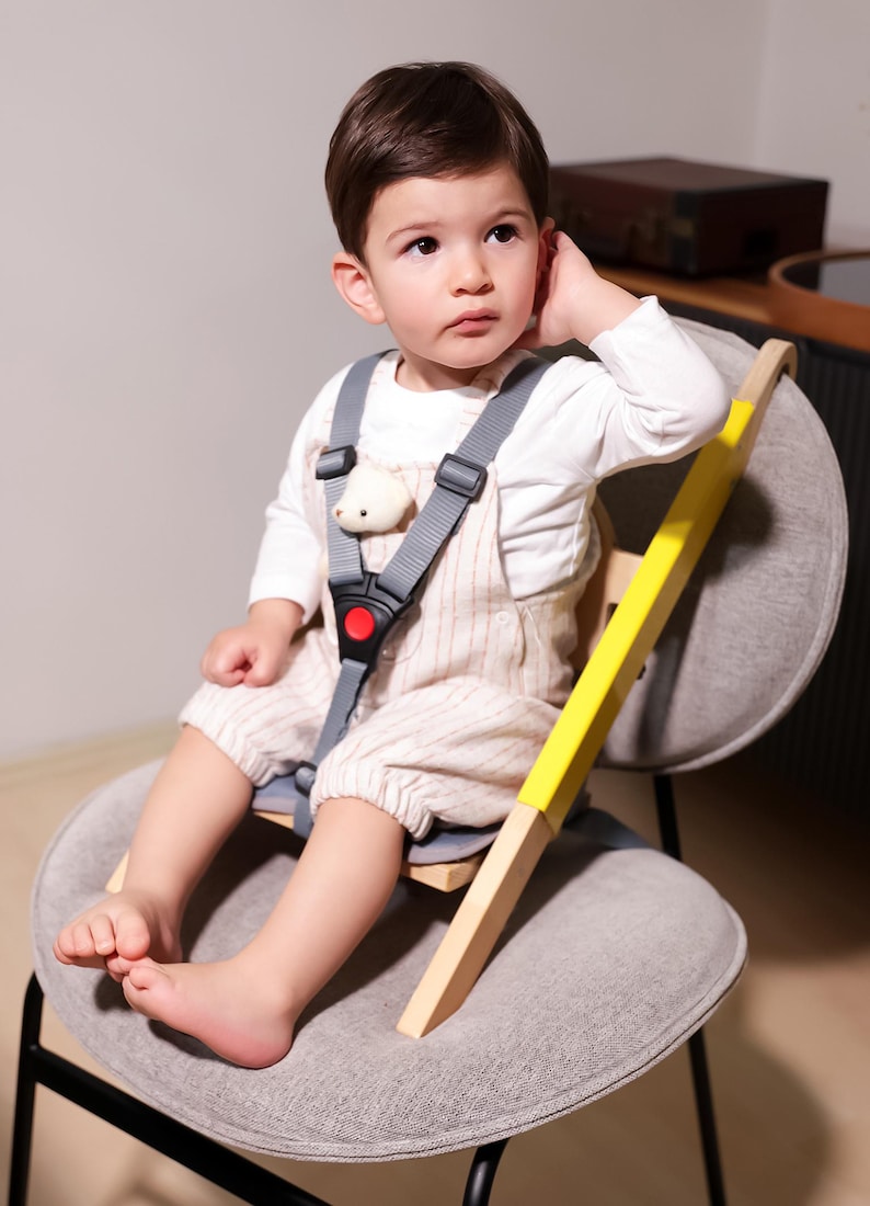 May include: A child is seated in a gray chair with a wooden high chair attachment. The high chair has a yellow accent and a gray safety harness with a red buckle. The child is wearing a white long-sleeve shirt and striped overalls.