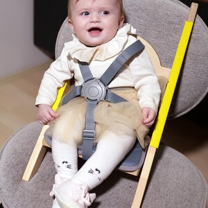 May include: A baby sits in a wooden high chair with a gray safety harness. The chair has yellow accents and is set on a gray upholstered chair. The baby wears a cream-colored top, a tulle skirt, and white socks with cat faces.