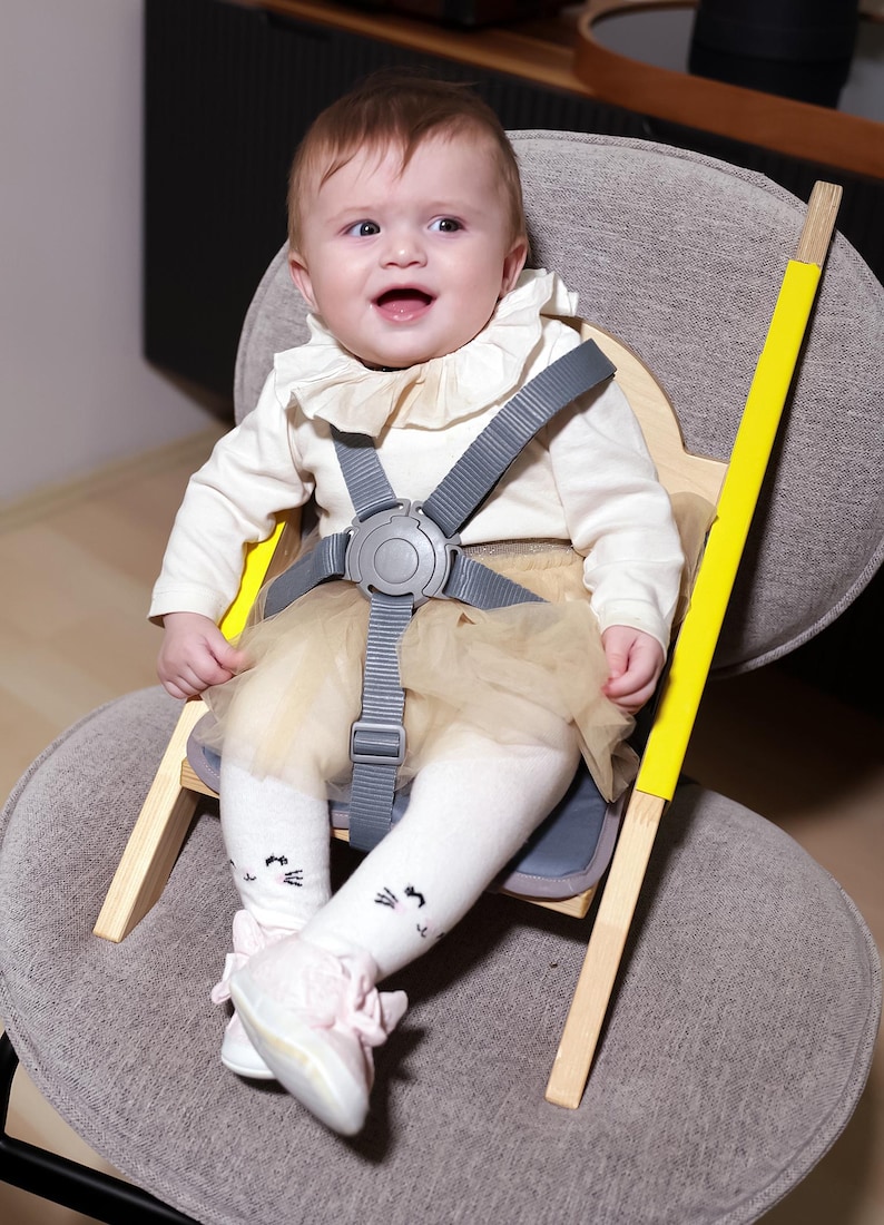May include: A baby sits in a wooden high chair with a gray safety harness. The chair has yellow accents and is set on a gray upholstered chair. The baby wears a cream-colored top, a tulle skirt, and white socks with cat faces.