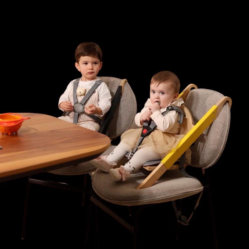 May include: Two babies seated in modern high chairs with gray fabric seats and wooden frames. One chair has a yellow accent. Both babies are secured with safety harnesses. A wooden table and an orange bowl are in the foreground.