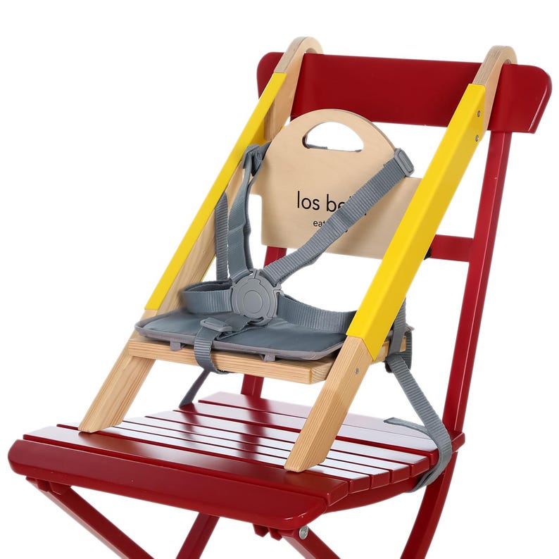 May include: A red wooden high chair with yellow accents and a gray safety harness. The chair has a natural wood seat and back, with the text "los be" printed on the backrest. The chair is designed for young children.