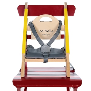 May include: A red and natural wood high chair with a gray safety harness. The chair has yellow accents and a wooden back with the text "los bella eat & joy". The seat is made of wood and has a red finish.
