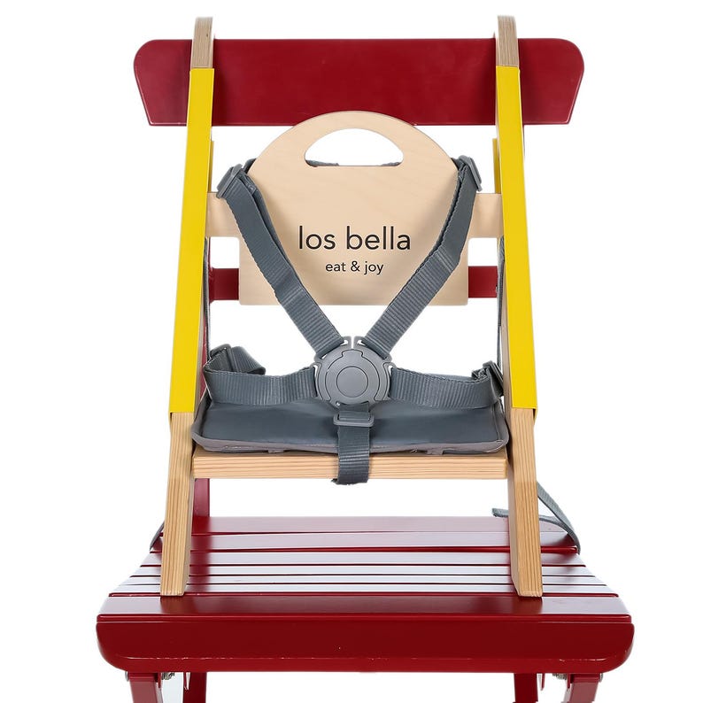 May include: A red and natural wood high chair with a gray safety harness. The chair has yellow accents and a wooden back with the text "los bella eat & joy". The seat is made of wood and has a red finish.
