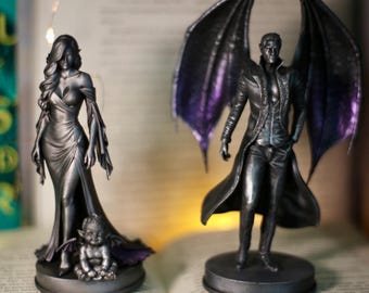 Puede incluir: Dos figuras metálicas oscuras sobre bases circulares. La figura de la izquierda es una mujer con un vestido largo y un bebé alado a sus pies. La figura de la derecha es un personaje masculino alado con un abrigo largo. Ambos tienen detalles morados.