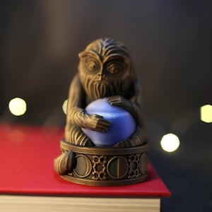 May include: A small, bronze-colored figurine of a monkey holding a blue orb. The monkey is sitting on a base with intricate details.