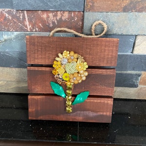 May include: A wooden wall hanging with a sunflower design made from colorful buttons and beads. The sunflower is on a brown wooden background with green leaves.