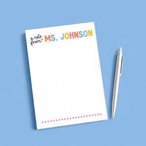 May include: A white rectangular notepad with the text "a note from MS. JOHNSON" printed in colorful letters at the top, a small pink heart icon, and a horizontal row of pink dots at the bottom, accompanied by a silver metallic ballpoint pen.
