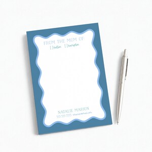 May include: A rectangular notepad with a blue border and a white wavy-edged center. The text reads FROM THE MOM OF, [ ] hatton [ ] hampton, NATALIE MARION, and 555 555 5555. A silver retractable ballpoint pen is positioned to the right of the notepad.