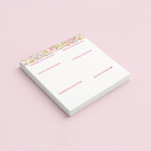 May include: A white notepad with pink text and a floral border. The notepad is divided into sections with prompts like "Verse of the Day," "Lift up to you," and "Thank you for." The notepad is on a light pink surface.