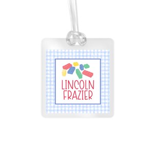 May include: A square luggage tag with a light blue and white gingham border. The center features a white square with a blue outline, a cluster of colorful plastic building blocks, and the red text LINCOLN FRAZIER. A clear plastic loop is attached.