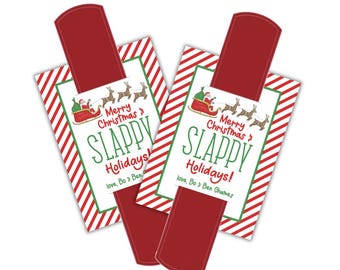 Könnte beinhalten: Zwei weihnachtliche Slap-Armband-Anhänger. Jeder Anhänger hat ein rotes Rechteck, das an einer weißen Karte mit rot-weißen Diagonalstreifen befestigt ist. Die Karte hat einen grünen Rand und trägt die Aufschrift "Merry Christmas & Slappy Holidays!" mit einer Grafik von Santa und Rentieren.